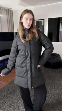 Černý péřový kabát s vysokým límcem, svislými švy a zapínáním na patentky. Má prošitou texturu a délku sahající ke kolenům.