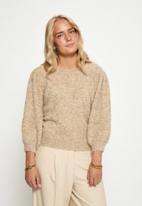 Bežani pleteni pulover s teksturiranim završetkom, okruglim izrezom i napuhanim rukavima, uparen s svijetlim naboranim hlačama.