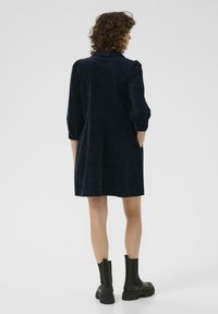 Donna dai capelli ricci di spalle, che indossa un vestito in velluto a coste di un blu scuro con maniche a tre quarti e stivali neri alla caviglia, su uno sfondo neutro.