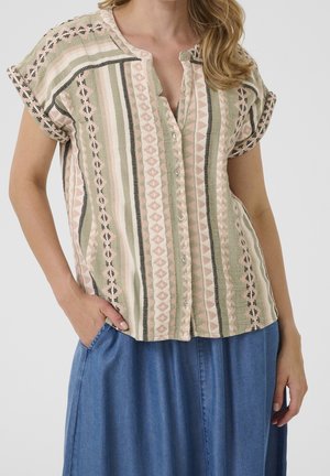 Kvinde iført kortærmet skjorte med knapper, med lodrette geometriske mønstre i pink, grøn og sort, og blå denimnederdel, hånd i lommen.