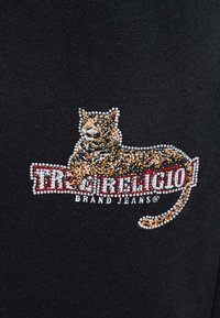 Melns audums ar stikla pērļu izšūtu leoparda dizainu, kas atrodas virs sarkanas un baltas "True Religion" zīmola džinsu logotipa teksta.