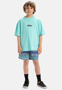 Camiseta de gran tamaño en color turquesa con un logotipo, combinada con pantalones cortos estampados en tonos azules y morados. Calcetines negros y zapatillas completan el atuendo.