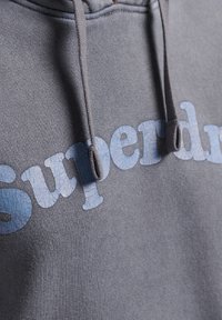 Felpa grigia con cappuccio caratterizzata da un logo "Superdry" di colore azzurro chiaro e texture, dotata di cappuccio regolabile con lacci grigi. Tessuto morbido con un design casual.