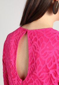 Ljusrosa spetsklänning med ett blommönster, med en nyckelhålsdesign bak och korta ärmar. Tyget verkar texturerat och lätt.