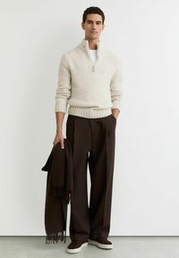 Bež pulover iz pletenine s polovičnim zadrgo na ovratniku, kombiniran s temno rjavimi širokimi hlačami in ujemajočo se ruto. Obleko dopolnjujejo bele superge.