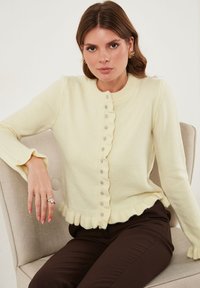 Vrouw met bruin haar, gekleed in een crèmekleurige gebuttonde trui met ruffles, donkere broek en gouden oorringen, zittend op een beige gestoffeerde stoel.