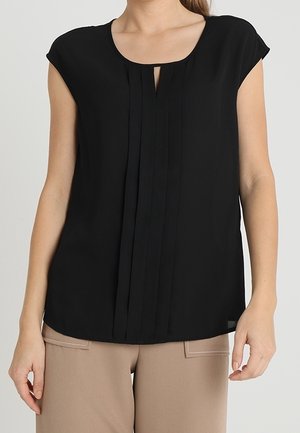 Black blouse with short sleeves, round neckline, and vertical pleats down the front. Smooth fabric with a relaxed fit. Paired with beige pants.