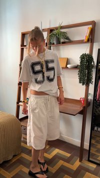 T-shirt branca com grande impressão preta "95", combinada com calções brancos largos e chinelos pretos. Acessórios dourados completam o look.