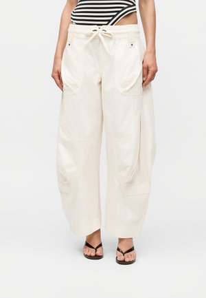 Γυναίκα που φοράει φαρδιά λευκά παντελόνια cargo με ρυθμιζόμενο κορδόνι στη μέση, μεγάλες μπροστινές τσέπες, μαύρα σαγιονάρες και ένα ριγωτό μπλουζάκι σε μαύρο και άσπρο.