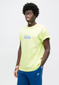 Κοντομάνικο μπλουζάκι σε νεον κίτρινο με γυρισμένα μανίκια, με κεντημένο το "café Swoosh" σε μπλε. Φορεμένο με μπλε φόρμα.