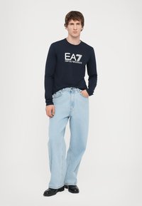 Marineblå langermet skjorte med hvit EA7-logo, kombinert med lyseblå vide jeans og svarte sko, stående foran en ensfarget bakgrunn.