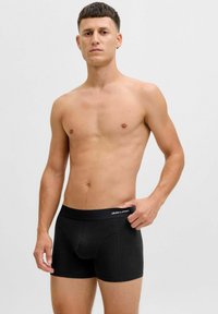 Black boxer briefs made from cotton with a soft waistband labeled "JACK & JONES"; model stands with a neutral expression against a plain background.