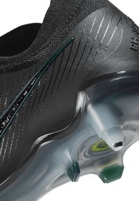 Scarpa sportiva nera con tomaia testurizzata, caratterizzata da linee riflettenti e una suola trasparente. Include borchie verdi e argentate per la trazione.