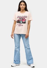 Roze katoenen t-shirt met een graphic van een rode Hummer met blauwe wielen en de tekst "Geen wegen nodig!" Vergezeld door lichtblauwe flared jeans.