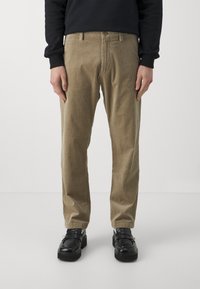 Osoba oblečená v černé mikině a béžových kordových kalhotách, stojící s rukama po stranách, doplněná černými slip-on botami.