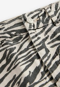 Gros plan sur un tissu en denim beige avec des rayures zébrées noires, avec un bouton en métal et des coutures visibles sur une ceinture.