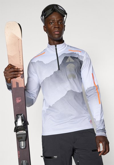 Camisa de esquí de manga larga gris claro con un gráfico de montaña, acentos naranjas y media cremallera. Gafas de esquí negras y esquís con fijaciones en la mano.