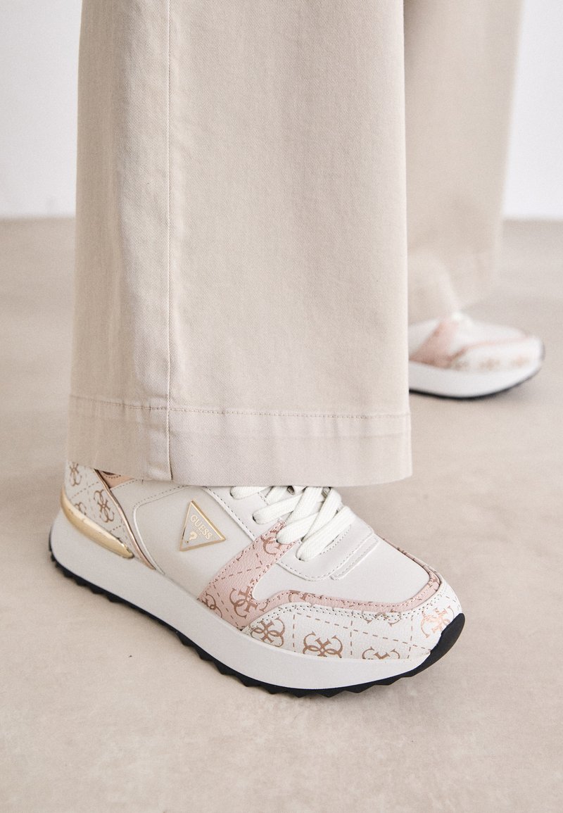 Sneaker bianco con accenti rosa e stampa monogram beige. Presenta un inserto dorato sul tallone, una suola testurizzata e lacci bianchi su una superficie piatta.