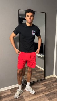 Νεαρός άνδρας με μαύρο κοντομάνικο με γραφικά, κόκκινα σορτς, λευκές κάλτσες και αθλητικά παπούτσια, στέκεται μπροστά σε καθρέφτη ολόκληρου σώματος σε δωμάτιο με ξύλινο πάτωμα.