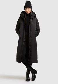 Sort lang puffjakke med hætte, som har en frontlynlås, trykknapper, side lommer og en glat tekstur. Kombineret med sorte leggings og støvler.