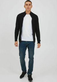 Cardigan noir à zip côtelé, porté sur un t-shirt blanc, associé à un jean bleu et des baskets noires. La texture est en tricot avec une coupe ajustée.