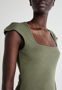 Top ajustado verde-azeitona com mangas curtas e estruturadas e decote quadrado. Tecido liso e elástico com um brilho subtil. Acento de colar dourado.