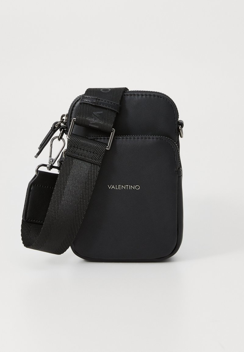 Svart crossbody-veske laget av glatt stoff, med glidelås, justerbar nylonstropp og foran et logo-detalj.