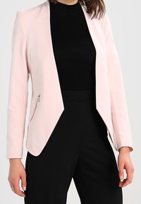 Ljusrosa kavaj med en skräddarsydd design, som har en ensam siddragkedja och långa ärmar, tillsammans med en svart ribbad topp.