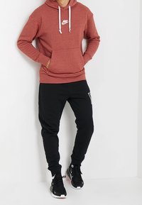 Man som bär en rödbrun Nike-hoodie, svarta Nike-joggingbyxor och svarta Nike-sneakers, som står mot en vit vägg.
