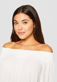 Witte off-shoulder blouse met smockhalslijn en vloeiende stof. Zachte textuur en een losse pasvorm, die de halslijn en schouders accentueert.