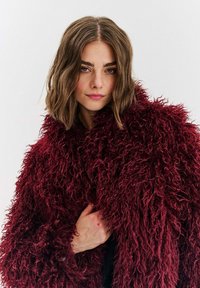 Žena s hnědými vlasy po ramena, která nosí velký, strukturovaný tmavě červený kožich z umělé kožešiny na jednobarevném bílém pozadí.