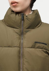 Person wearing an olive green padded jacket with a high collar and a visible zipper, featuring the "PEGADOR" logo on the chest.
