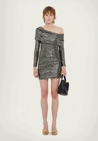 Fekete flitteres mini ruha, vállra csúszó fazonnal és hosszú ujjakkal. A modell fekete kézitáskát tart, arany szandálban van. Simasága kiemelkedő.