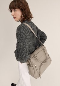 Borsa tote in pelle color cuoio con molteplici scomparti con zip, design ricco di funzionalità e tracolla regolabile, abbinata a un maglione a maglia grigio scuro.