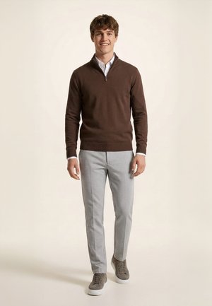 Homme souriant, marchant vers l’avant portant un pull marron à fermeture éclair quart, une chemise blanche, un pantalon gris clair et des baskets marron sur fond uni.