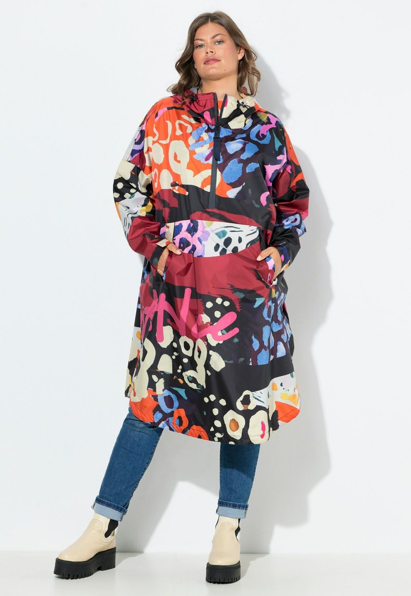 Femme portant une veste surdimensionnée colorée à motifs avec poche avant, un jean bleu et des bottes crèmes épaisses, se tenant contre un fond blanc.