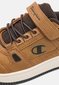 Zapatillas de cuero marrón con una lengüeta de tela texturizada en marrón oscuro, suela de goma negra y costuras naranjas. Cuenta con una correa de velcro y un logo.