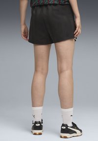 Πόδια και κάτω μέρος κορμού ενός ατόμου που φοράει μαύρες σορτς, λευκές κάλτσες Puma και μαύρα αθλητικά παπούτσια Puma σε γκρίζο φόντο.