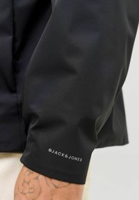 Veste noire avec une finition mate lisse, vue en gros plan du poignet affichant le logo Jack & Jones en gris clair.