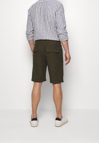 Shorts verde oliva con un tessuto testurizzato, dotati di due tasche posteriori con alette. Abbinati a una camicia a righe con maniche lunghe e scarpe da ginnastica.