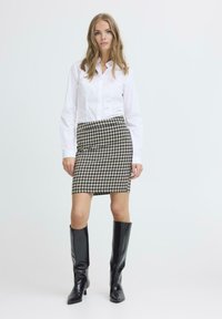 Weißes Hemd, Rock im Hahnentrittmuster in Schwarz und Beige, knielange schwarze Lederstiefel, stehend vor einem schlichten Hintergrund.