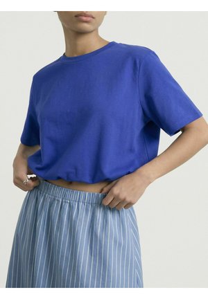 Kvinde iført ensfarvet blå t-shirt med korte ærmer, som er stukket ned i en højhæklet blå og hvidstribet nederdel med elastik i taljen.