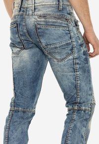 Verblasste blaue Jeans aus Denim mit kontrastierendem Nähen, Taschenakzenten und einem Reißverschlussdetail auf der Rückseite. Das Markenlabel ist eingeprägt.