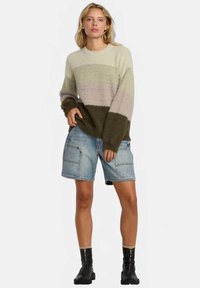 Vaalea nainen, jolla on päällään raidallinen pörröinen neule, polvitason farkkushortsit ja mustat nilkkurit, seisoo yksinkertaista taustaa vasten.