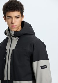 Giacca a vento nera e grigia con due tasche anteriori con zip, tessuto testurizzato e logo sulla manica sinistra. Presenta un colletto alto e un cappuccio regolabile.