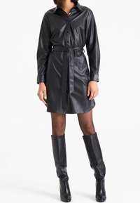 Abito camicia in finta pelle nera con bottone anteriore, maniche lunghe, vita definita con fascia, e stivali neri in pelle al ginocchio. Texture liscia, design classico.