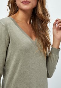 Sweter w kolorze oliwkowej zieleni z dekoltem w serek, wykonany z miękkiego dzianinowego materiału, z prążkowanym kołnierzem i mankietami, o lekko dopasowanym kroju i gładkiej fakturze.