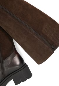 Brown suede boot with a black rubber sole, featuring a side zipper and smooth leather accents. The design includes a textured panel for detail.