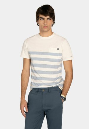 Giovane uomo che indossa una camicia a maniche corte a righe bianche e azzurre con una tasca sul petto, pantaloni blu scuro e un orologio da polso.