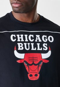 Črna bombažna majica s kratkimi rokavi z napisom "CHICAGO BULLS" v belem. Prikazuje rdečo glavo bika z črnimi podrobnostmi in belimi rogovji.
